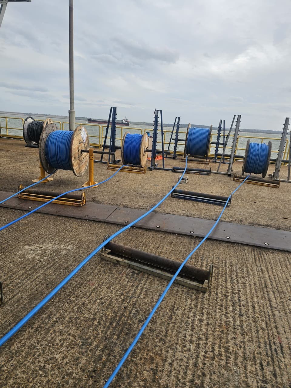 Cable drums set up on stands and rollers on the terminal jetty, with the estuary and a passing ship beyond