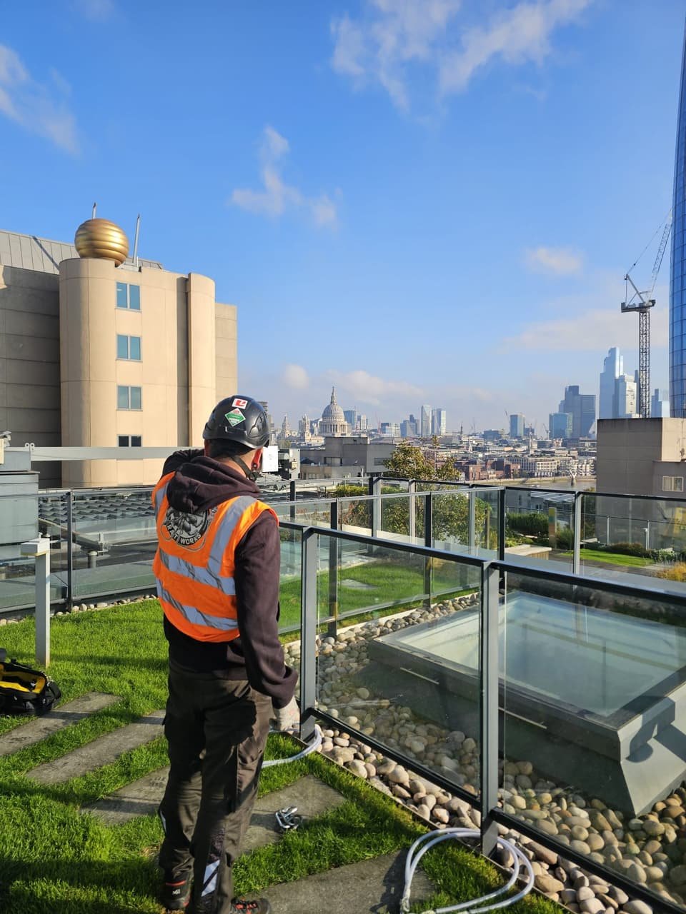RAT Work technician in hi-vis on a London roof garden beside the glazing, with the City skyline behind