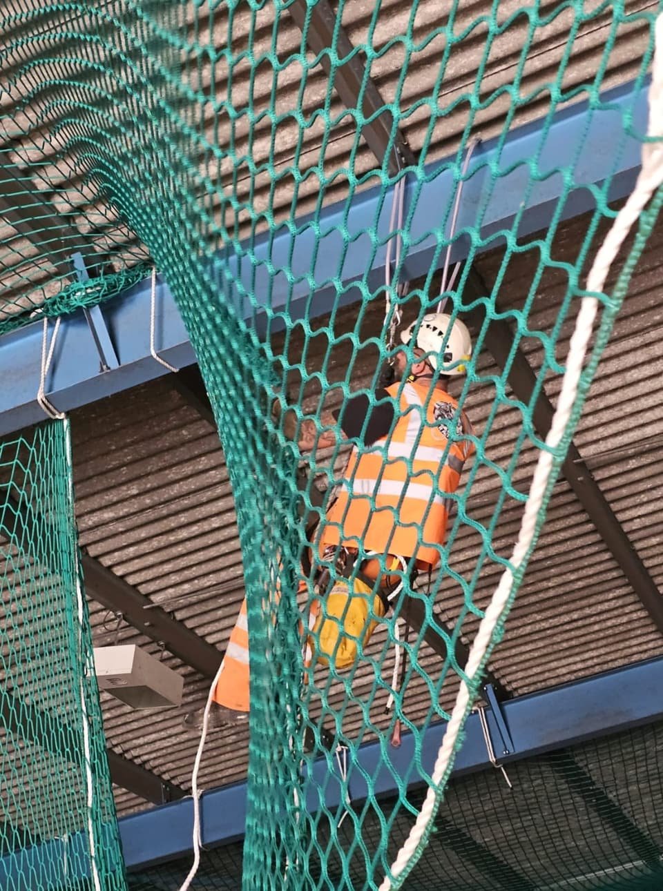 Technician inside a safety net rigged beneath a roof structure, seen from below