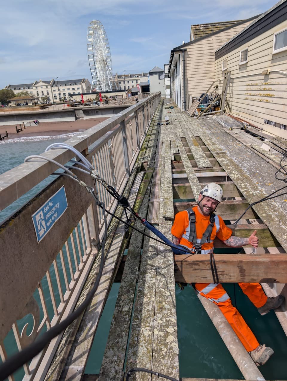 Technician on rope in the open decking of a pier during timber repairs, with the seafront behind